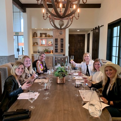 Six women seated at a long dining table, holding wine glasses and smiling.