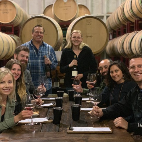 Group of people smiling and holding wine glasses in a winery with barrels.