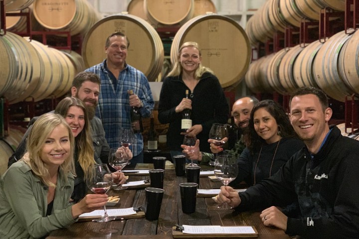 Group of people smiling and holding wine glasses in a winery with barrels.