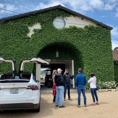 People gathered near a white car with open doors in front of an ivy-covered building.