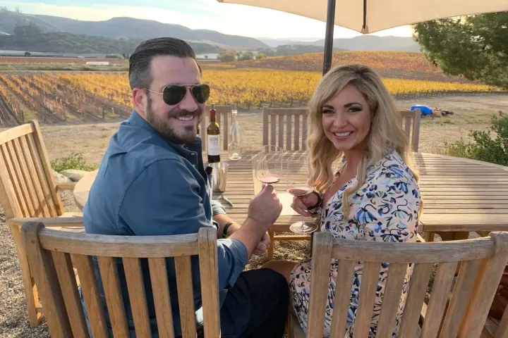 Couple sitting at a winery table, toasting with wine glasses, vineyards in background.