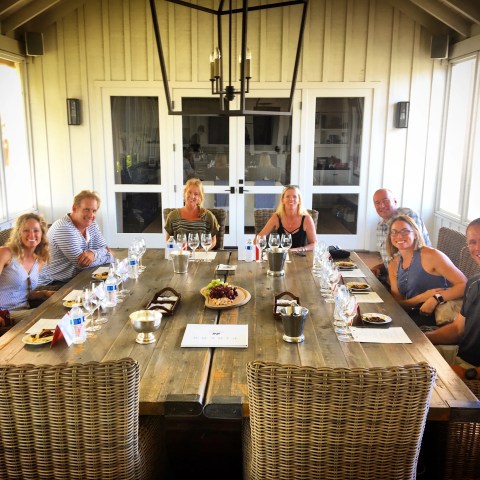 Eight people smiling at a dining table set with wine glasses and plates in a well-lit room.