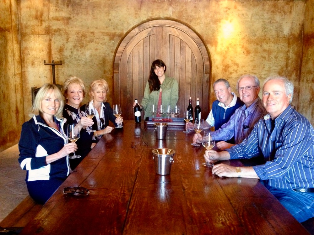 Seven people sitting at a wooden table with wine glasses in a rustic room.