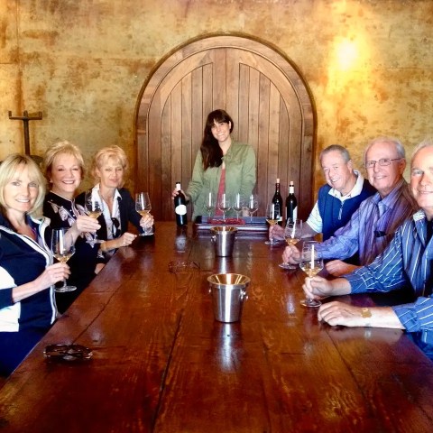 Seven people seated at a wooden table, holding wine glasses in a rustic room with a wooden door.