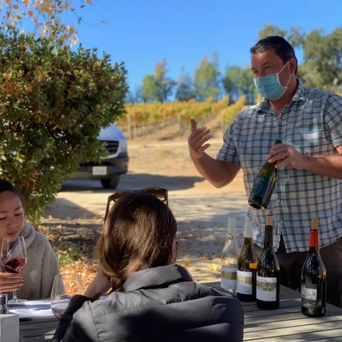 Masked man presenting wine to seated women outdoors near vineyard.