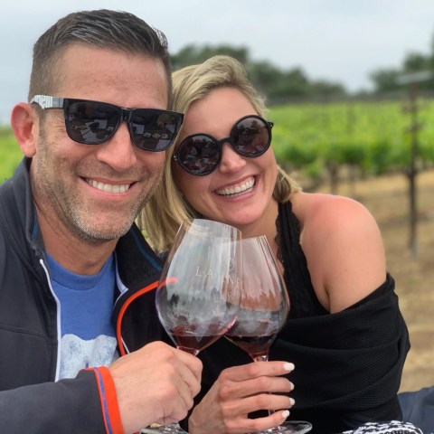 Smiling couple in sunglasses toasting with wine in a vineyard.