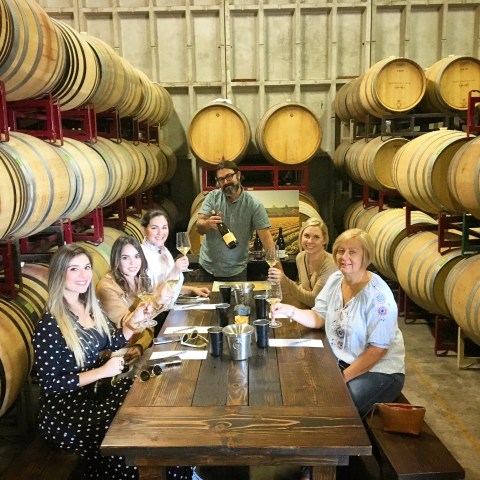 Group of people tasting wine at a wooden table in a wine cellar with barrels in the background.