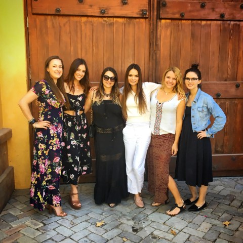 Six women in colorful outfits standing in front of a large wooden door.