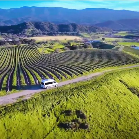 Aerial view of a white van on a path by green fields and mountains in the background.