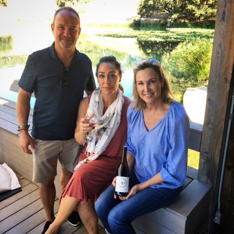 Three people sitting on a deck by a pond, one holding a wine bottle, another with a glass.