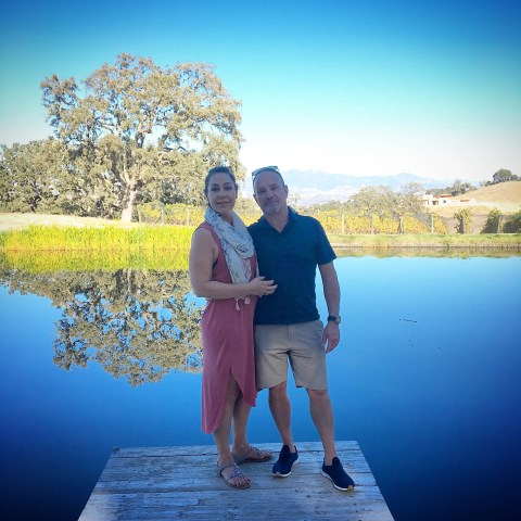 Two people standing on a wooden dock by a lake with trees and mountains in the background.