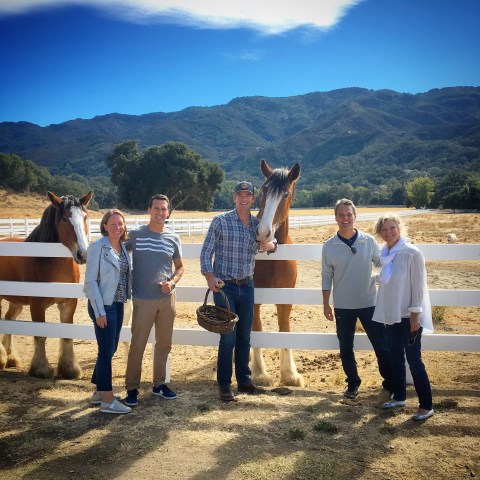 Five people standing by fences with two horses and mountains in the background.