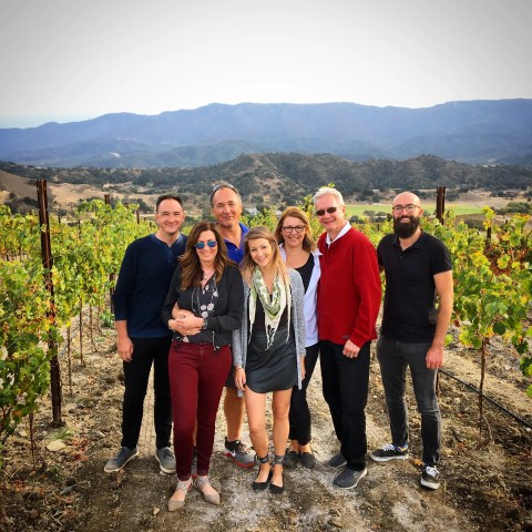 Group of seven people posing in a vineyard with mountains in the background.