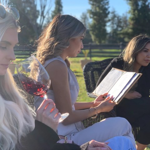 Three women sitting outdoors, one holding a glass of wine, another reading a menu.