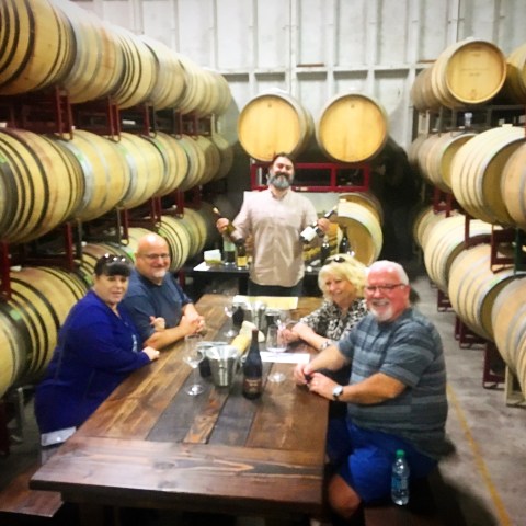 Five people sitting at a table in a wine cellar with barrels around them.