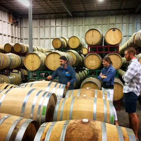 Three people inspecting stacked wooden barrels in a warehouse.
