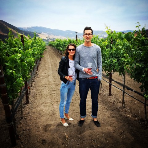 Couple in vineyard holding wine glasses, with green grapevines and hills in the background.