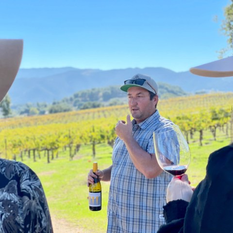 Man in vineyard talking, holding wine bottle; people in hats holding wine glasses listen.