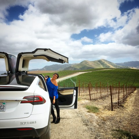 Woman in blue top leans against a white car with open falcon doors, in a vineyard with hills in the background.