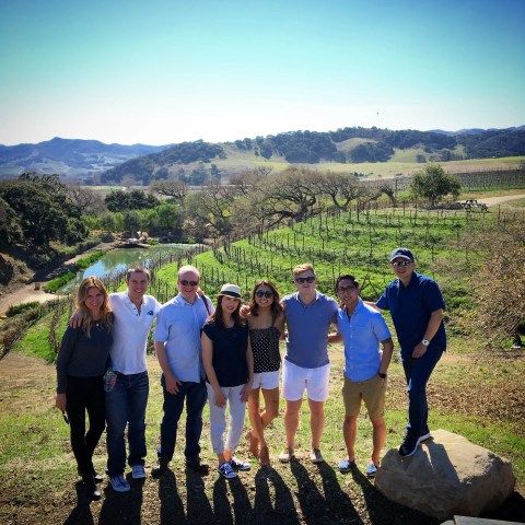 Group of people standing in front of a scenic vineyard landscape under a clear blue sky.