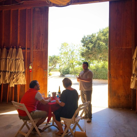 Three people sitting and talking inside a barn with open doors and trees visible outside.
