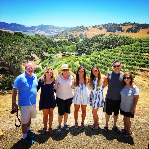 Group of seven people posing in front of vineyards with hills and clear sky in the background.