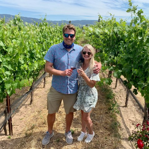 Couple in sunglasses holding wine glasses, standing in a vineyard on a sunny day.