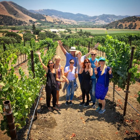 Group of people posing in a vineyard with mountains in the background under a clear blue sky.