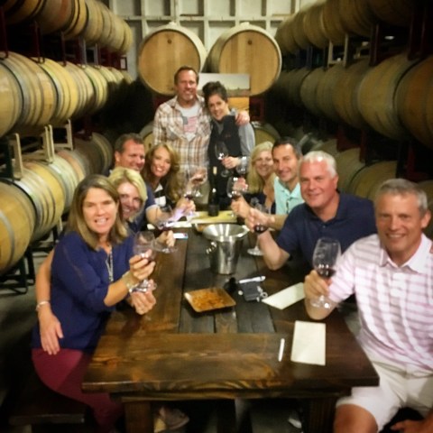 Group of people sitting at a winery table with barrels, holding wine glasses.