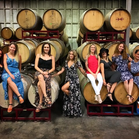 Eight women in dresses pose on wine barrels in a cellar.