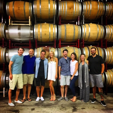 Group of eight people standing in front of stacked wine barrels in a cellar.