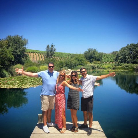 Four people with arms outstretched stand on a dock by a pond with lush greenery and vineyard in the background.