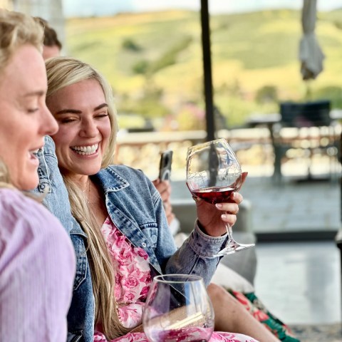 Two women smiling and holding wine glasses in an outdoor setting.