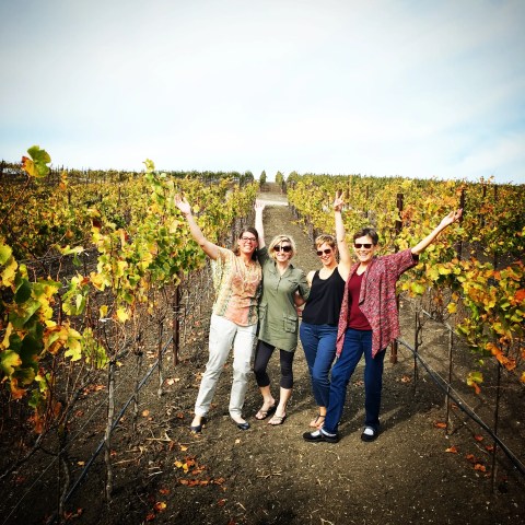 Four women posing happily in a vineyard with autumnal leaves and raised arms.