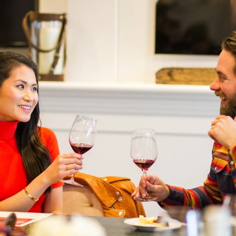 Two people smiling and toasting with red wine glasses at a table.