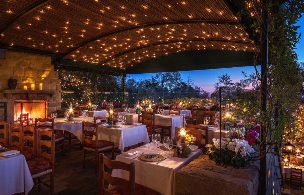 Outdoor restaurant setting with string lights, covered patio, lit fireplace, and white tablecloths at sunset.