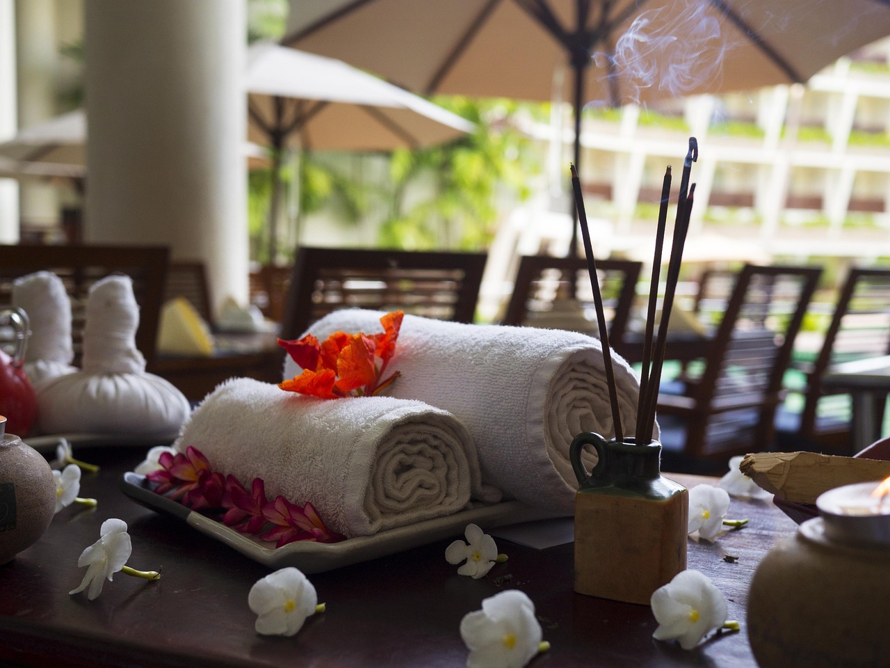 Rolled towels with flowers on a spa table, incense sticks burning, outdoor setting with umbrellas.
