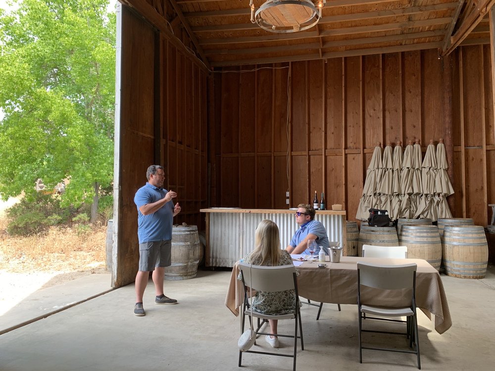 Man speaking to two seated people in a barn with wine barrels and bottles nearby.