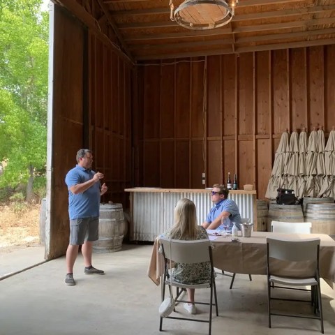 Man speaking to two seated people in a rustic wooden barn with wine barrels.