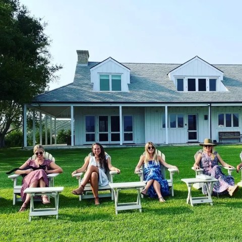 Six women in sundresses sit on white chairs in front of a large white house with a green lawn.