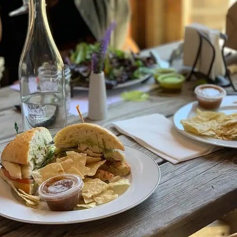 Plates with sandwiches, chips, and sauce on a wooden table with water bottle and wine glasses.