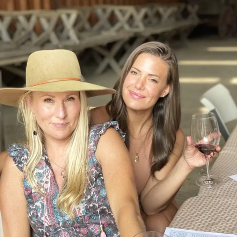 Two women smiling, seated at a table with wine glasses and papers.