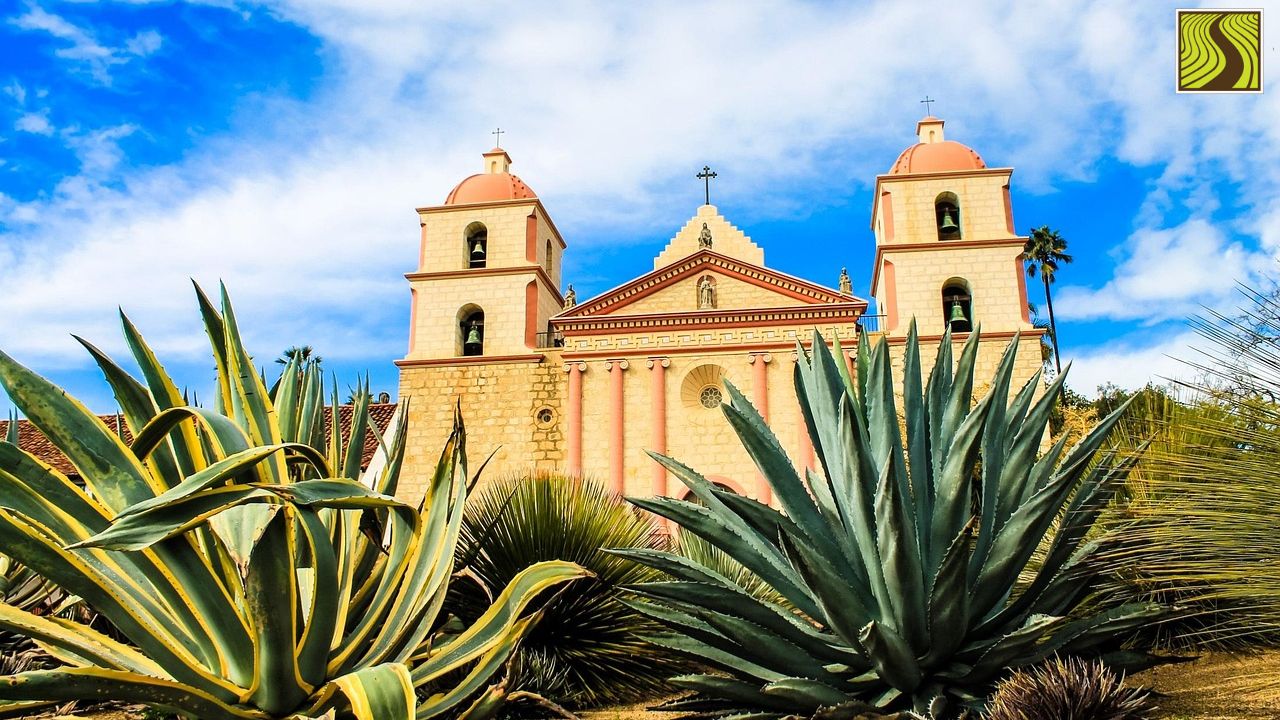Historic church with two towers seen through large agave plants under a blue sky.
