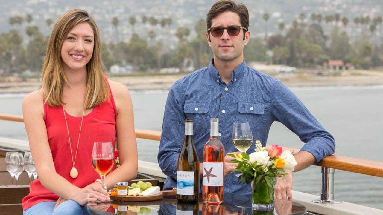 A woman and man dining on a boat with wine, cheese, and flowers, with a scenic coastline in the background.