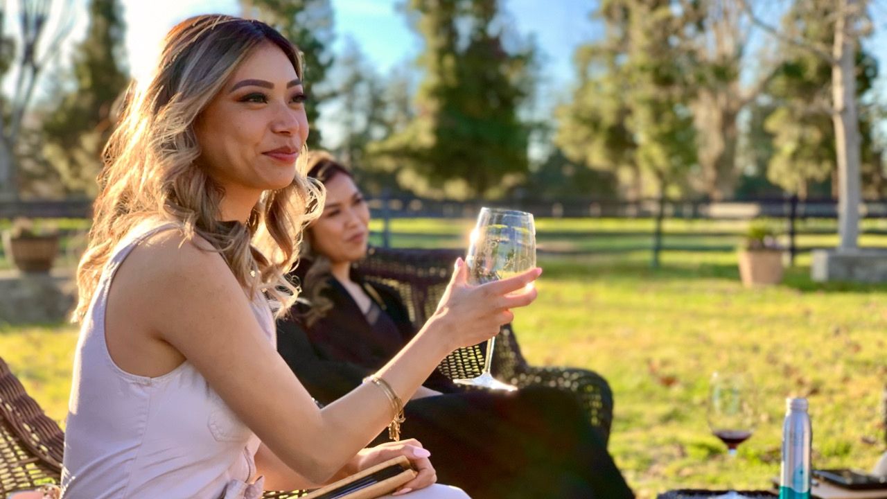 Two women sitting outdoors, one holding a glass, with trees in the background.