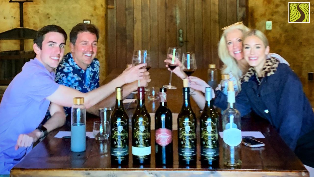 Four people toasting with wine glasses at a table with wine bottles.