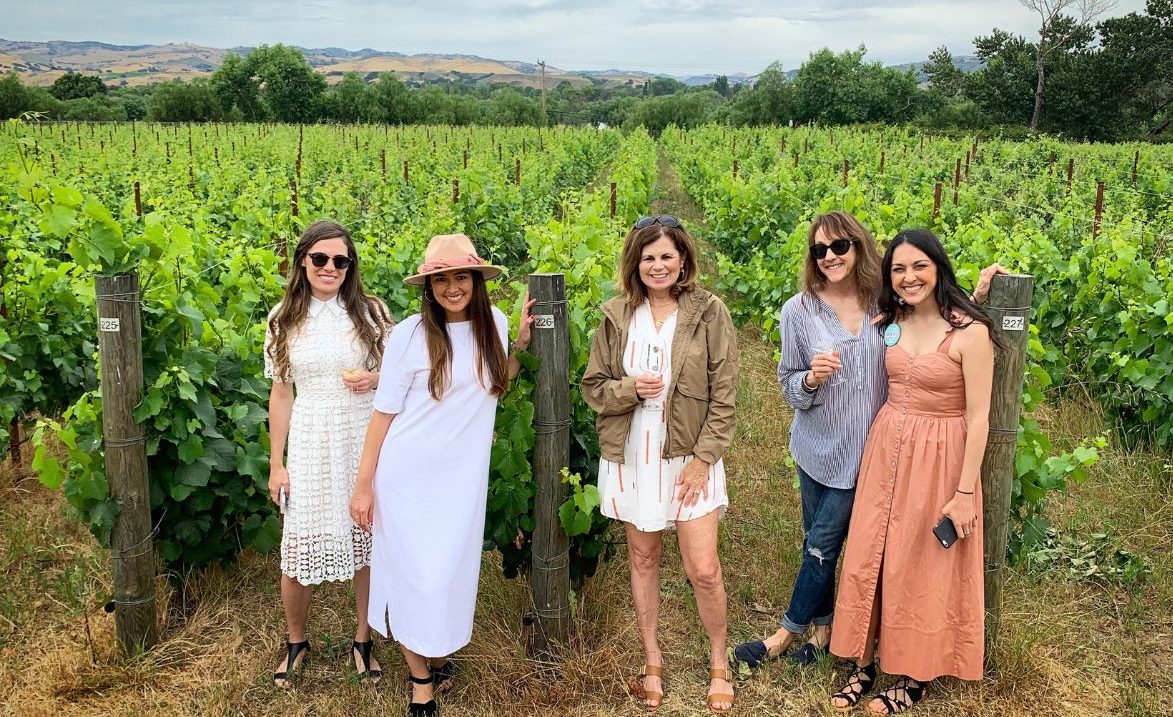 Five women posing in a vineyard with a scenic background.