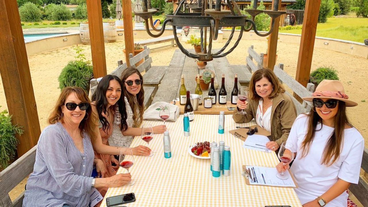 Five women smiling, seated outdoors at a table with wine glasses, bottles, and fruit.