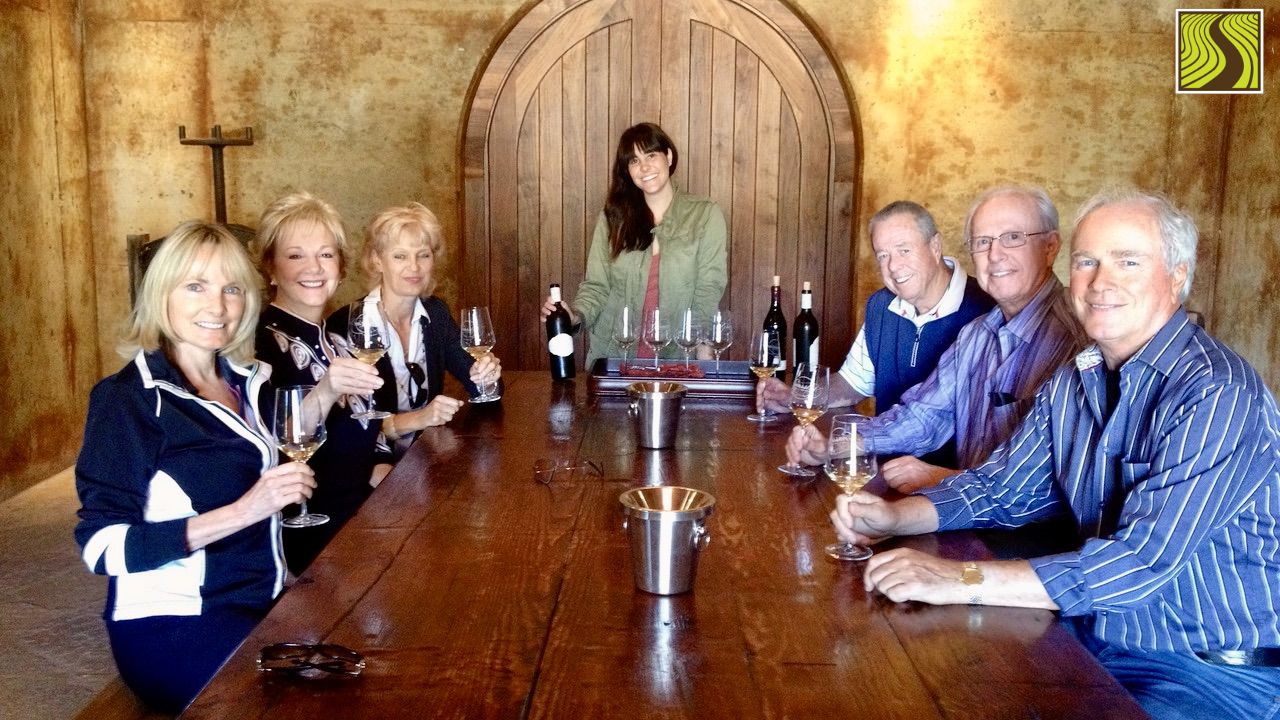Group of people seated around a table indoors at a wine farm, tasting wine.