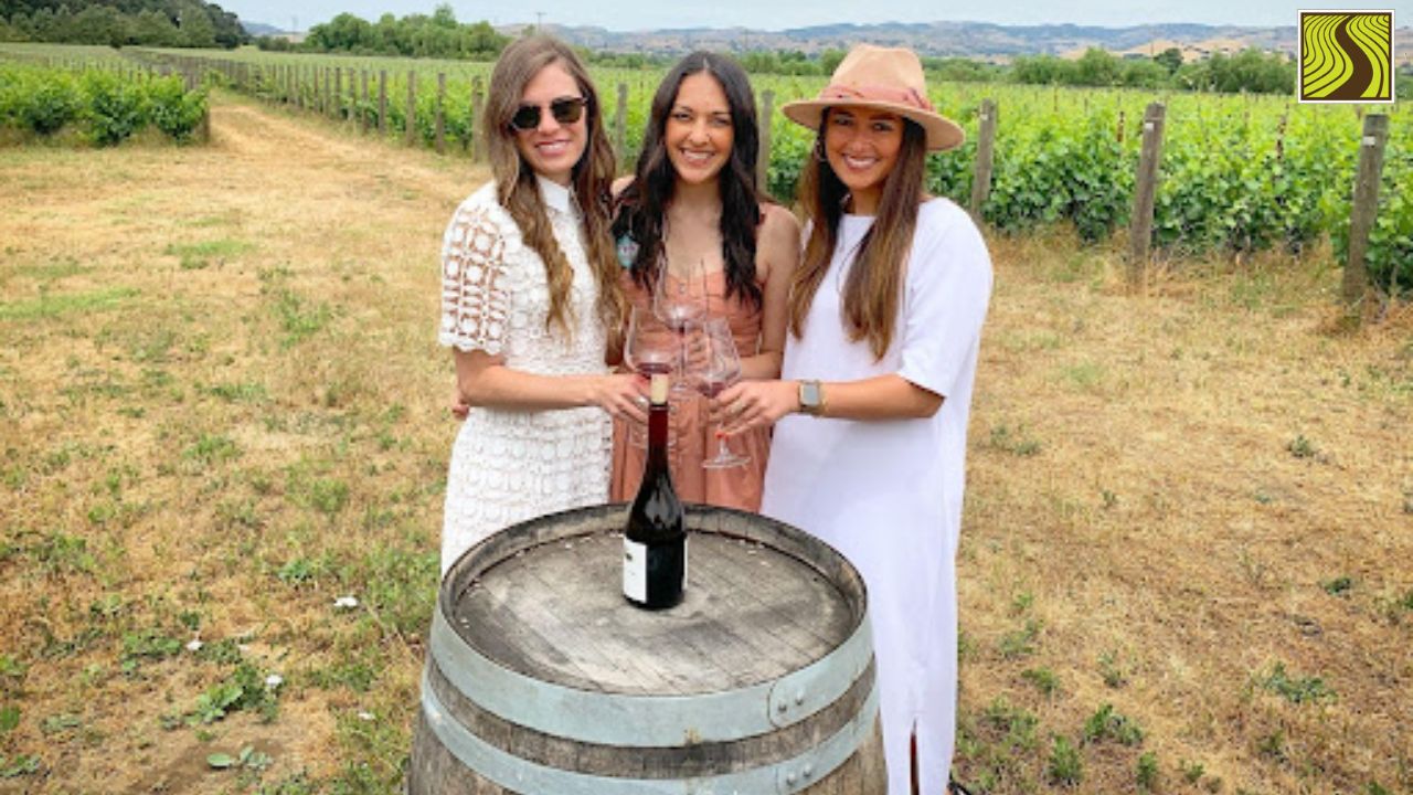Three women standing behind a wine barrel in summer attire in a vineyard